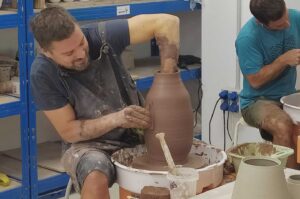 Un hombre da forma a un jarrón de arcilla en un taller de cerámica con torno, rodeados de herramientas para su elaboración.