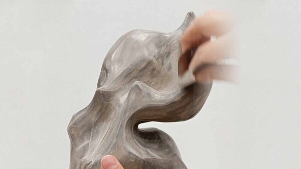Una mano perfila con precisión una escultura abstracta orgánica de tono gris terroso, suavizando sus formas fluidas y textura ligeramente áspera
